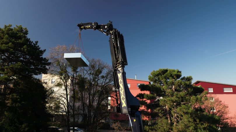 Ein Kran hebt ein Teichbecken über Gebäude und Bäume vor klarem blauen Himmel.